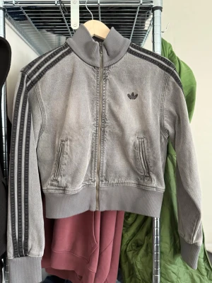 Grå jeansjacka från Adidas - Säljer en grå ziptröja från Adidas Originals med klassiska svarta ränder längs ärmarna och Adidas-logga på bröstet. Tröjan har hög krage, muddar vid ärmslut och nederkant samt två fickor framtill. Perfekt för en sportig och avslappnad stil.
