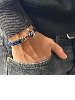 Blått armband med ankardetalj - Snyggt armband i blått rep med en silverfärgad ankare som lås. Armbandet har en modern och stilren look, perfekt för dig som gillar marina detaljer. Passar både till jeans och t-shirt eller mer uppklätt. Materialet är textil och metall.