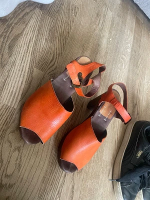 Orange läderskor med träklack - Säljer ett par snygga orange sandaler i läder med öppen tå och justerbart spänne runt ankeln. Skorna har en träfärgad klack och en stilren design som passar perfekt till sommaren. Klassisk svensk stil med robust känsla. Är lite slitna i spännet och framtill. Lite missfärgningar och fläckar. Lädret har sjunkit ihop lite som man ser på sista bilden. 
