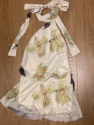 Vit blommig halterneckklänning - Säljer en vit halterneckklänning med blommigt mönster i grönt, beige och ljusa toner. Klänningen har knyt i nacken och är ärmlös med en flowy passform. Den har även inbyggda shorts under. Väldigt fin och skön på sommaren, perfekt inför påsk och midsommar!