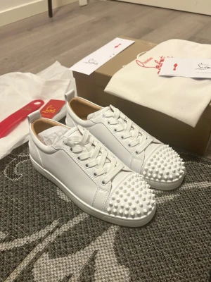 Vita Christian Louboutin sneakers med nitar - Säljer ett par vita sneakers från Christian Louboutin med coola vita nitar på tån. Skorna har klassisk låg profil, vita skosnören och är gjorda i slätt läder. Perfekta för dig som vill sticka ut med en edgy och lyxig stil.