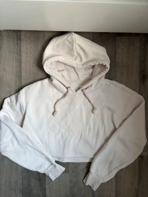 Ljusbeige cropped hoodie från Lounge - Säljer en ljusbeige cropped hoodie från Lounge med broderad logga framtill. Hoodien har huva med dragsko och långa ärmar. Tillverkad i mjukt bomullsmaterial som känns riktigt skönt mot huden. Perfekt för en chill och avslappnad stil. Använd 1 gång