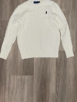 Vit kabelstickad tröja Polo Ralph Lauren - Kabelstickad vit tröja från Polo Ralph Lauren med klassisk rund hals och svart broderad logga på bröstet. Tröjan har långa ärmar och ribbade muddar vid ärmslut och nederkant. Perfekt för lager på lager och stilrena outfits.