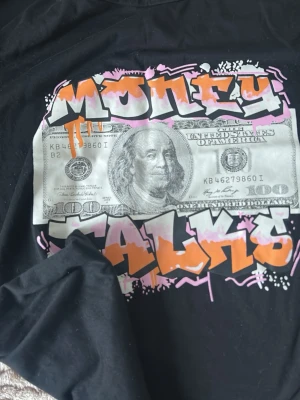 Svart Money Talks t-shirt - Cool svart t-shirt med stort tryck av en dollarsedel och graffiti-texten 'Money Talks' i vitt, orange och rosa på framsidan. T-shirten har rund hals och korta ärmar. Perfekt för dig som gillar streetwear och statement-plagg.