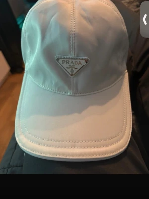 Vit keps från Prada med metallogo - Snygg vit keps  med den ikoniska trekantiga metalloggan framtill. Kepsen har böjd skärm och är tillverkad i ett glansigt material som ger en exklusiv känsla. Perfekt accessoar för att lyfta din outfit.