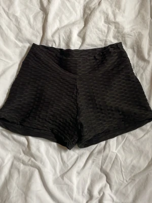 Svarta ribbade shorts med scrunch - Svarta shorts med ribbad struktur och scrunch-detalj bak. De har en tight passform och är tillverkade i ett stretchigt syntetmaterial som sitter skönt på kroppen. 