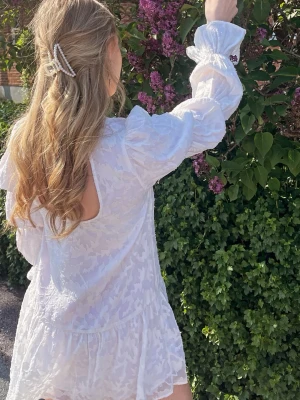 Vit kortklänning med öppen rygg - Supersöt vit kortklänning med volangkant och puffiga långärmar. Klänningen har ett subtilt blommigt mönster och en snygg öppen rygg. Perfekt för varma dagar och somriga tillfällen. Materialet känns lätt och luftigt.