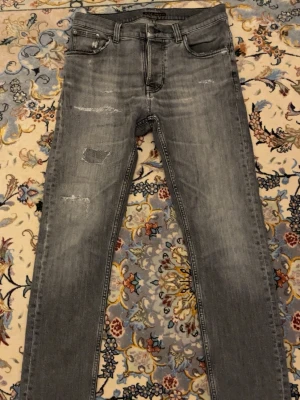 Grå slitna jeans från Nudie Jeans - Säljer ett par gråa slim fit jeans från Nudie Jeans med slitningar och distressed detaljer på framsidan. Klassisk femficksmodell med svarta broderade detaljer på bakfickorna. Jeansen har normal midja och är tillverkade i stretchigt denimtyg. Modellen är Grim Tim och denna färg slutades sälja för flera år sedan, storlek 31/32