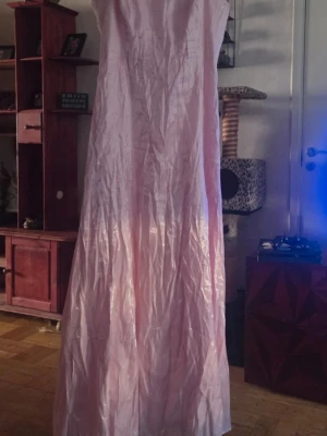 Ljusrosa långklänning i satin - Elegant ljusrosa långklänning i glansigt satinmaterial. Klänningen är axelbandslös och har en klassisk, rak siluett som faller mjukt mot golvet. Perfekt för speciella tillfällen när du vill känna dig extra fin.