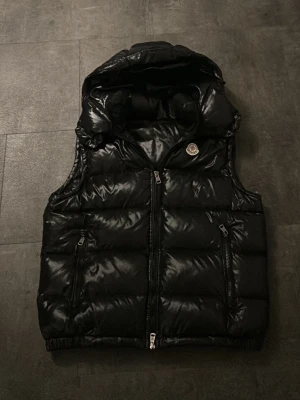 Monclerväst - Snygg svart dunväst från Moncler med glansig finish och avtagbar huva. Västen har två dragkedjeförsedda fickor och klassisk Moncler-logga på bröstet.