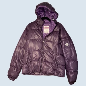 Lila pufferjacka från champion - Snygg lila pufferjacka från champion med glansig finish och stor huva. Jackan har två snedställda fickor framtill, dragkedja och tryckknappar vid ärmsluten. Märkets logga finns på ärmen. Perfekt för kalla vinterdagar.