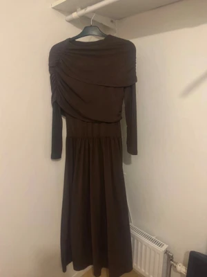 Brun långklänning med drapering - Snygg brun långklänning med långa ärmar och draperad detalj över axlar och överkropp. Klänningen har en mjuk, flytande passform och är perfekt för dig som gillar en elegant och stilren look. Tillverkad i ett stretchigt material som känns bekvämt.