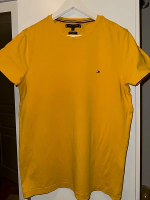 Gul t-shirt från Tommy Hilfiger - Snygg gul t-shirt från Tommy Hilfiger i ekologisk bomull. Slim fit-modell med korta ärmar och rund halsringning. Liten broderad logga på bröstet och diskret randig detalj på insidan av kragen. Perfekt för dig som gillar stilrena plagg med en färgklick.