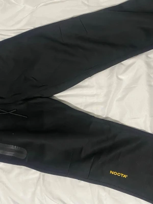 Svarta NOCTA mjukisbyxor - Säljer ett par svarta mjukisbyxor från NOCTA med gul logga på benet och diskreta detaljer som 'AWR' och 'CL' broderat i gult. Byxorna har dragsko i midjan och fickor på sidorna. Perfekta för en chill och stilren look.