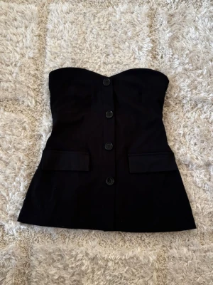 Svart axelbandslös tubtopp med knappar - Snygg svart axelbandslös tubtopp med stora knappar framtill och två dekorativa fickor. Toppen har en stilren och elegant look, perfekt att styla med jeans eller kjol. Materialet känns mjukt och har en lätt struktur.