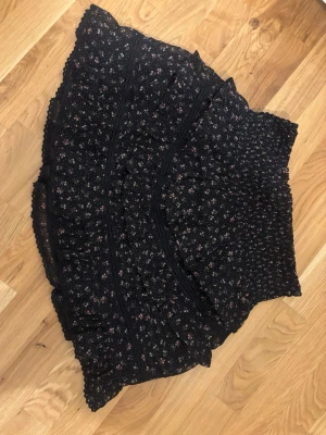 Blommig volangkjol från Neo Noir - Supersöt kortkjol från Neo Noir i svart med små blommor i rosa och vitt. Kjolen har volangdetaljer och smock i midjan för en bekväm passform. Perfekt att styla med en enkel topp eller stickad tröja. Helt ny❤️