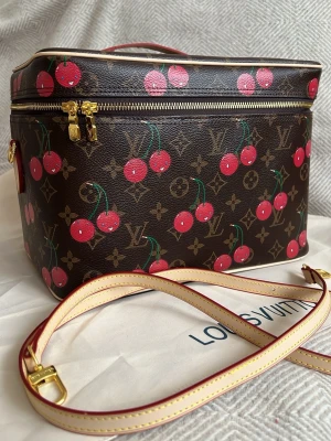 Louis Vuitton Cherry Monogram handväska - Unik handväska från Louis Vuitton med klassiskt brunt monogrammönster och färgglada körsbärsdetaljer. Väskan har guldfärgade metalldetaljer, avtagbar axelrem i ljust läder med röda kanter och dragkedja upptill. Perfekt för dig som vill sticka ut med en ikonisk design. Den största storleken i sminkväska 