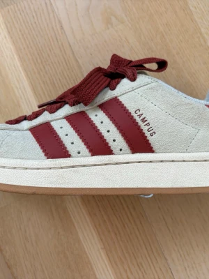 Adidas Campus sneakers beige/röd mocka - Säljer ett par Adidas Campus sneakers i beige mocka med klassiska röda ränder och breda röda snören. Skorna har en låg profil och gummisula. Perfekta för dig som gillar retrostil och vill sticka ut med snygga detaljer. Använda fåtal gånger. 