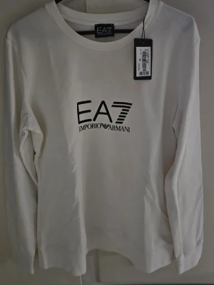 Vit långärmad topp från Emporio Armani - Snygg vit långärmad topp från Emporio Armani EA7 med svart logotyptryck framtill och på ryggen. Klassisk rund halsringning och stilren design som passar till det mesta. Tillverkad i mjuk bomull för en bekväm känsla.