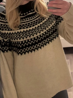 Beige stickad tröja med svart mönster - Säljer en beige stickad tröja med svart och vitt nordiskt mönster över bröstet och axlarna. Tröjan har hög krage, långa ärmar och ribbade muddar. Perfekt för kalla dagar och riktigt mysig att bära.