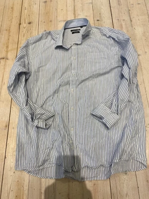 Randig skjorta från Madison Avenue - Klassisk vit skjorta med tunna blå ränder från Madison Avenue. Skjortan är tillverkad i 100% bomull, har lång ärm, knappar framtill och en tidlös krage. Perfekt för en clean och stilren look.