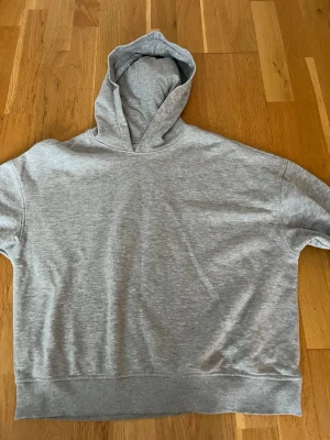 Grå basic hoodie med huva - En enkel och stilren grå hoodie med huva och ribbade muddar vid ärmsluten. Hoodien har en avslappnad passform och är tillverkad i mjukt bomullsmaterial. Perfekt för en chill och casual look.