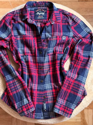 Röd och marinblå rutig skjorta Superdry - Klassisk rutig skjorta från Superdry i rött och marinblått med vita detaljer. Skjortan har långa ärmar, knappar framtill och två bröstfickor. Tillverkad i mjuk bomull, perfekt för en avslappnad och cool stil.