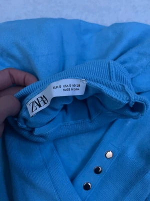 Ljusblå stickad tröja från Zara - Trendig ljusblå stickad tröja från Zara med långa ärmar och snygga guldfärgade knappar längs ärmsluten. Tröjan har ribbade muddar och är tillverkad i ett mjukt, tunt material som passar perfekt till olika outfits. Använt 1 gång 