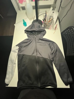 Grå och svart hoodie från H&M Sport - Snygg hoodie från H&M Sport i grått och svart med hel dragkedja framtill och huva. Tillverkad i mjuk polyester, perfekt för chill dagar eller träning. Långärmad modell med två fickor framtill och sportig look.