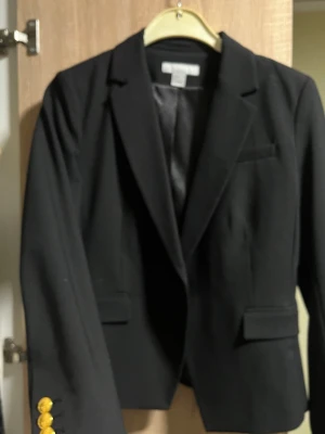 Svart kavaj från H&M med guldknappar - Stilren svart kavaj från H&M i storlek 38. Klassisk modell med två fickor fram och snygga guldknappar på ärmsluten. Kavajen har slät finish och är helfodrad med svart tyg. Perfekt för en trendig och uppklädd look.