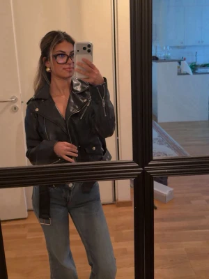 Svart klassisk skinnjacka - Säljer en svart skinnjacka i klassisk bikerstil med breda slag, dragkedjor och bälte i midjan. Jackan har coola detaljer som axelklaffar och silverfärgade knappar. Perfekt för dig som vill ha en tidlös och edgy look.