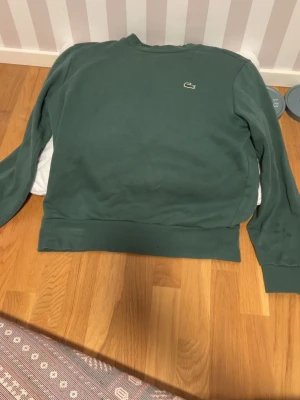 Grön sweatshirt från Lacoste - Mörkgrön sweatshirt från Lacoste med klassisk rund halsringning och den ikoniska krokodilloggan broderad på bröstet. Tröjan har ribbade muddar vid ärmslut och nederkant, perfekt för en avslappnad och stilren look.