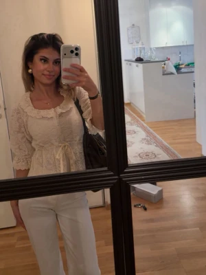 Söt spetsblus med rosett i midjan - Romantisk cremefärgad spetsblus med trekvartsärm och volangkant vid halsen. Blusen har en markerad midja med knytband och fina spetsdetaljer över hela plagget. Perfekt för dig som gillar en feminin och vintageinspirerad stil.