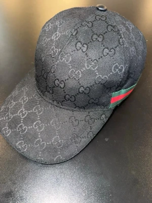 Gucci keps - Snygg svart keps från Gucci med det klassiska GG-monogrammet över hela kepsen. På sidan finns en grön och röd rand som ger en ikonisk touch. Kepsen är tillverkad i tyg och har en böjd skärm. Perfekt för dig som gillar exklusiva accessoarer.