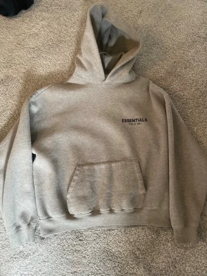 Grå Essentials Fear of God hoodie - Säljer en grå hoodie från Essentials Fear of God med stor huva och känguruficka. Tryck med 'ESSENTIALS' på bröstet och ryggen. Oversized passform och mjukt material, perfekt för en chill och trendig look. Bara att fråga om ni undrar över något!