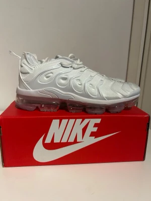 Nike Air Vapormax Plus vita sneakers - Nike Air Vapormax Plus sneakers i helvitt med futuristisk design och genomskinlig sula. Ovandelen är i textil och syntet med distinkta vågformade detaljer och klassisk Nike-logga på plösen. Skorna har snörning och dragflik bak för enkel påtagning.