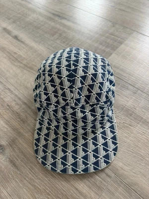 Lacoste keps  - Säljer en snygg blå keps från Lacoste med vitt geometriskt mönster och klassisk böjd skärm. Kepsen har metallspänne på sidan och är tillverkad i ett mjukt tygmaterial. Perfekt för dig som vill sticka ut med en unik accessoar.