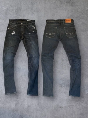 Vintage Selvedge replay anbass  - Mörkblå Replay Anbass jeans med snygga slitningar och patchdetaljer framtill. Klassisk femficksmodell med kontrastsömmar och logotyp på bakfickan. Jeansen har en rak passform och är gjorda i slitstarkt denimtyg. Perfekt för dig som gillar en cool och avslappnad stil.