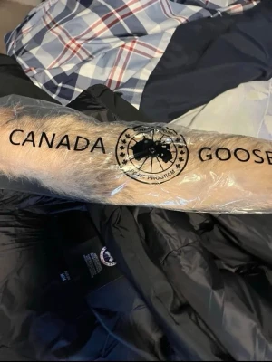 Svart dunjacka från Canada Goose - Säljer en svart dunjacka från Canada Goose med avtagbar huva och beige fuskpälsdetalj. Jackan har klassisk patch på ärmen, dragkedja och knappar framtill samt flera fickor. Perfekt för kalla vinterdagar och riktigt varm.