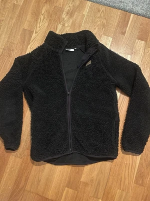 Svart teddyjacka med dragkedja - Mysig svart teddyjacka med hel dragkedja framtill och hög krage. Storleken är 170 men jag har haft den och jag är 182 men skulle rekommendera ca 170-175 då den satt ganska litet på mig. Perfekt för kalla dagar när du vill vara både varm och stilren.