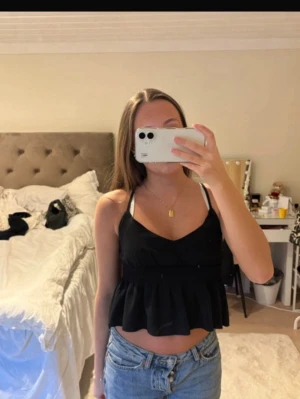 Svart croppad linnetopp - Snygg svart croppad linnetopp med smala axelband och volang nedtill. Toppen har en smockad midja som ger en skön och figurnära passform. Perfekt till jeans eller kjol för en trendig look.