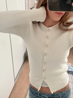 Vit kofta med pärlknappar - Söt vit kofta med långa ärmar och dekorativa pärlknappar framtill. Koftan har ribbade muddar vid hals, ärmslut och nederkant samt en figurnära passform. Perfekt att styla med jeans eller kjol för en trendig look.