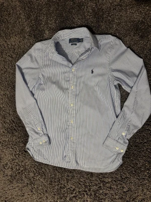 Randig skjorta från Polo Ralph Lauren - Klassisk blåvit randig skjorta från Polo Ralph Lauren i slim fit-modell. Den är i riktigt fint skick utan några defekter. Storleken är 18 ungefär xs och passar mellan 160-170. Tveka inte på att höra av er😊