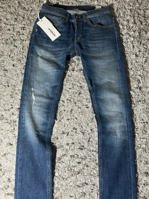Blå slim fit jeans från Dondup George - Säljer ett par klassiska blå jeans från Dondup med raka ben och snygga slitningar på låret. Jeansen har fem fickor, normal passform och är tillverkade i mjukt bomullsmaterial. Perfekta för en avslappnad och stilren look. Modell george. Helt nya med schyssta slitningar. Längd: 102cm midja: 38 cm