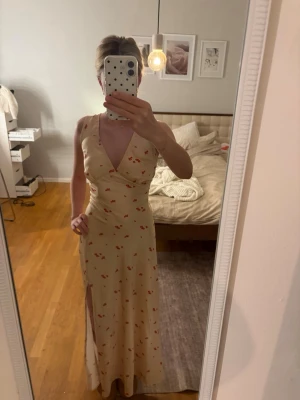 Satin klänning  - Gul/champange färgad långklänning med blommigt mönster i rött och omlottdesign. Klänningen har v-ringning, är ärmlös och har en slits på sidan. Perfekt för sommarens alla tillfällen. Finns ett band som knytes i ryggen