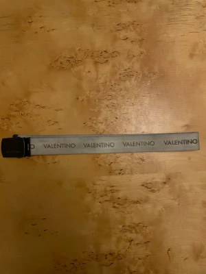  Valentino bälte  - Snyggt grått bälte från Valentino med svart spänne och tydlig VALENTINO-logga längs hela bandet. Perfekt accessoar för att lyfta din outfit med en exklusiv touch. Bältet har en modern och stilren design.