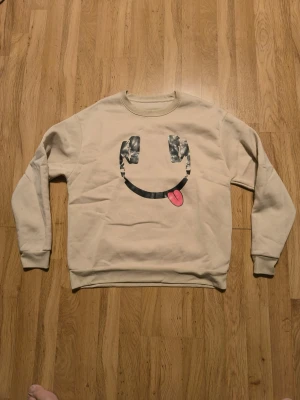Beige sweatshirt med smiley-tryck - Cool beige sweatshirt med ett stort smiley-tryck på framsidan där ögonen är i camomönster och tungan är rosa. Tröjan har rund halsringning, långa ärmar och ribbade muddar vid ärmslut och nederkant. Perfekt för en avslappnad och trendig look.