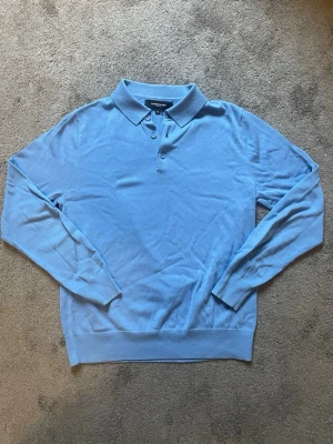 Lawrence Grey blå långärmad polotröja - Snygg blå långärmad polotröja från Lawrence Grey med klassisk krage och tre knappar framtill. Tröjan är tillverkad i ett mjukt stickat material och har ribbade muddar vid ärmslut och nederkant. Perfekt för dig som gillar stilrena plagg. 