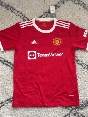 Manchester United matchtröja Adidas - Röd Manchester United matchtröja från Adidas med vita detaljer, klubbmärke på bröstet och TeamViewer som sponsortryck. Tröjan har korta ärmar, rund halsringning och är tillverkad i lätt, ventilerande material perfekt för fotboll.