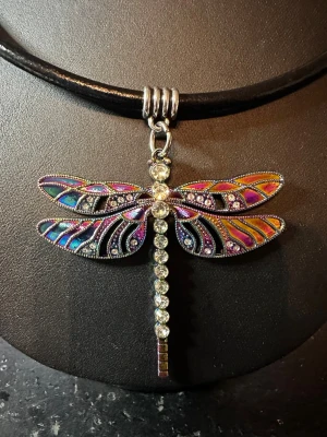 Halsband med trollslända och pärlor - Unikt halsband med svart läderrem och en stor trollsländehänge i multifärg. Vingarna har färgglada detaljer i lila, blått och orange samt gnistrande stenar. Kroppen är dekorerad med klara pärlor som ger extra bling. Perfekt för dig som vill sticka ut med accessoarerna.